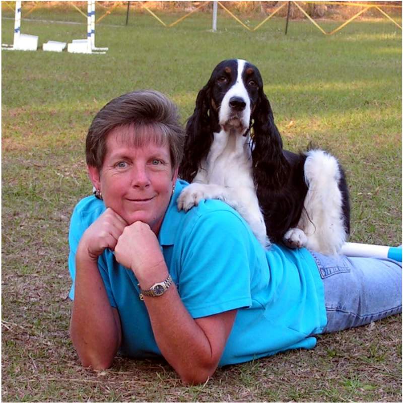 Lady with short brown hair wearing jeans and a turquoise shirt is lying on prone with chin on hands. Black and white springer spaniel, Luci sitting on lady’s back looking at the camera
