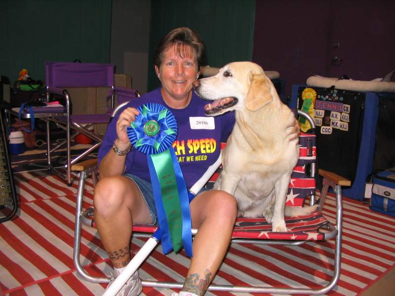 The same lady, in pink shirt kneeling beside black and white springer spaniel, Luci flanked by two female agility one holding red, white and blue rosette award ribbon and white PVC autograph bar.